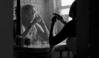 Black and white image of playwright and activist Leeza Jessie Peterson reflected in a mirror, with a brick wall and window visible behind her.